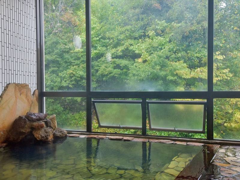 【大浴場】男湯からの眺め。外に緑を眺め、清流のせせらぎが聞こえる、自慢のお風呂。広がる自然の風景とお湯の温かさに、身も心もゆだね、ゆっくりとおくつろぎください